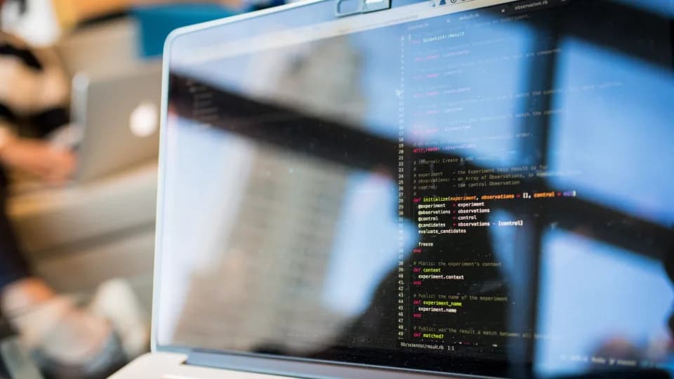 A laptop on a desk by a window showing lines of code on a dark editor, out-of-focus city buildings behind