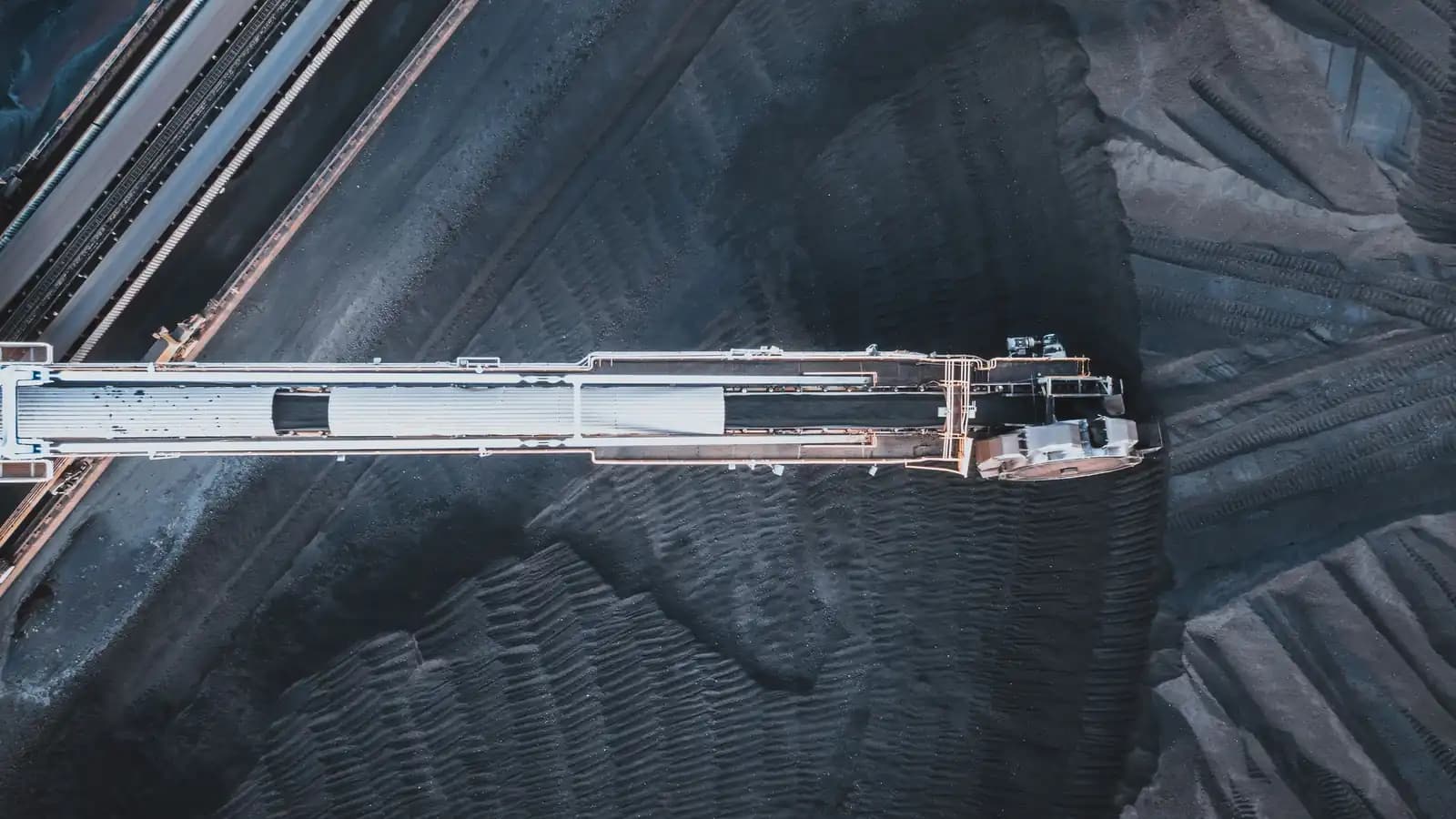 Overhead view of a long white conveyor bridge crossing a dark stockyard of bulk material, with rail lines running along one edge