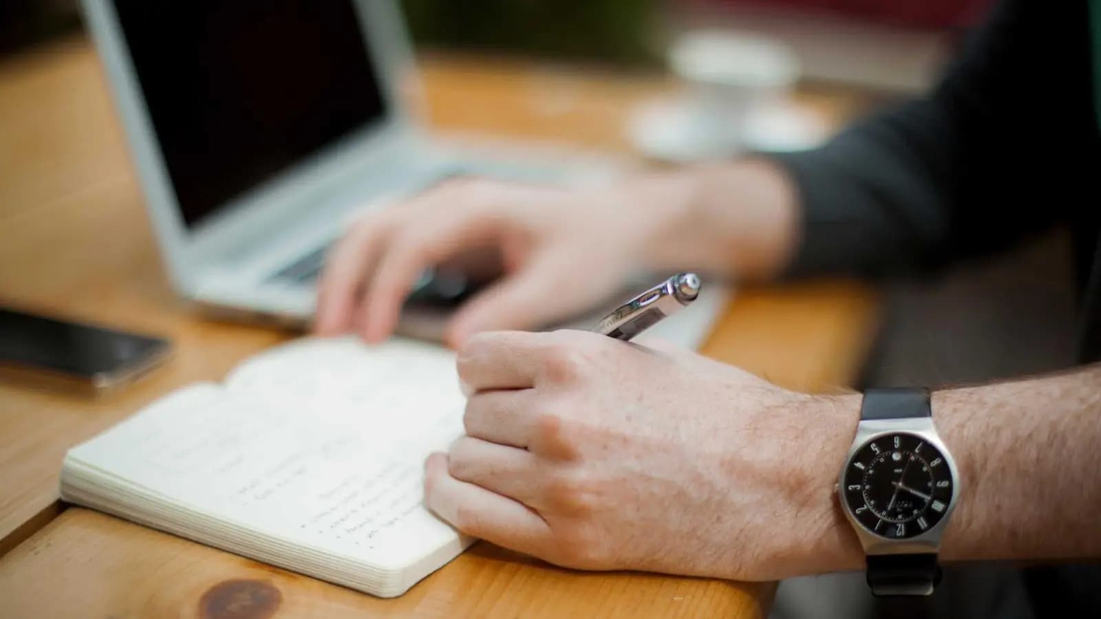 Close-up of a person at a wooden desk writing in a small notebook beside an open laptop, a wristwatch on their arm