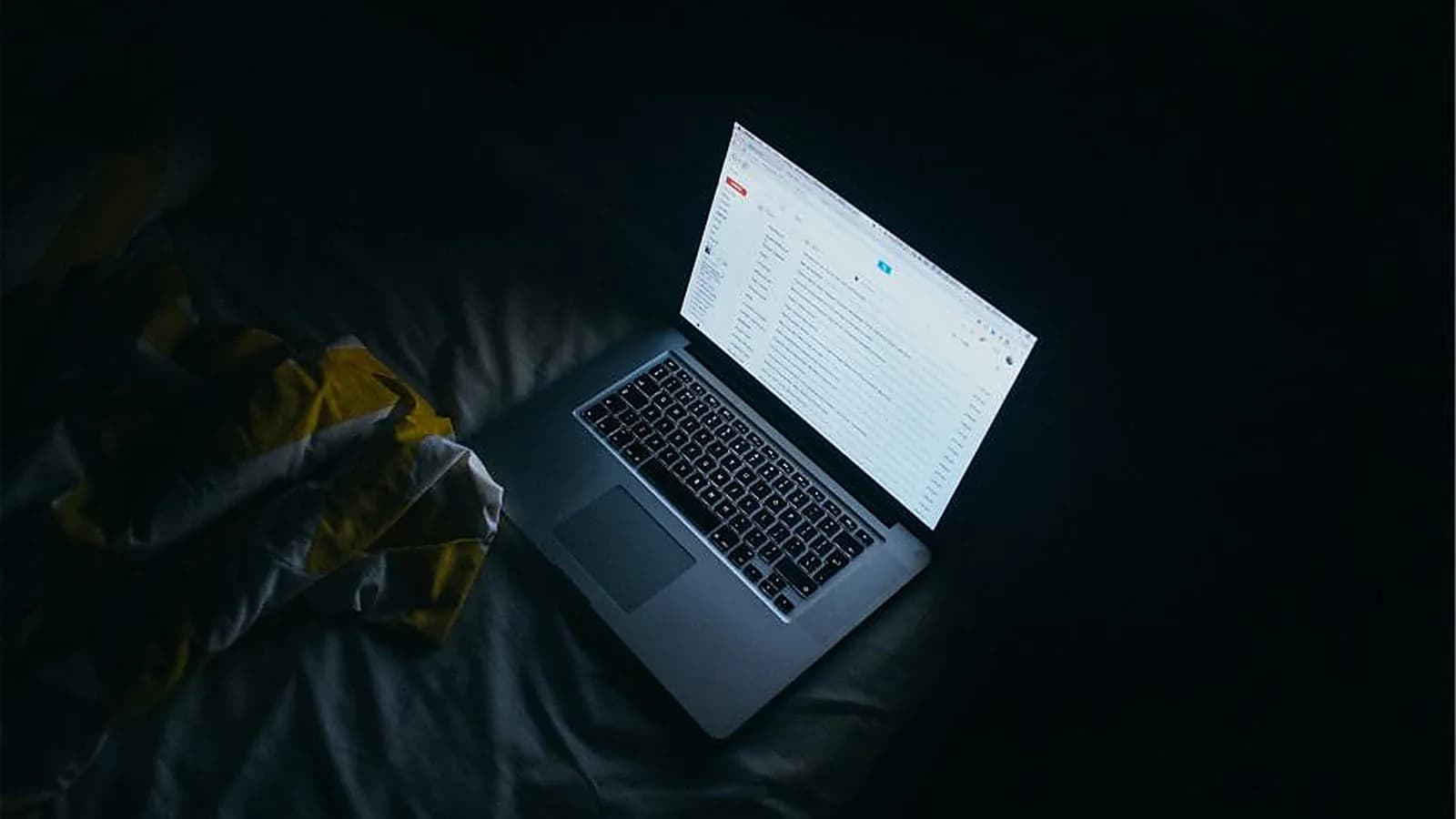 Photograph: an open laptop in a darkened room, its screen the only light, showing a long list of messages in an email client