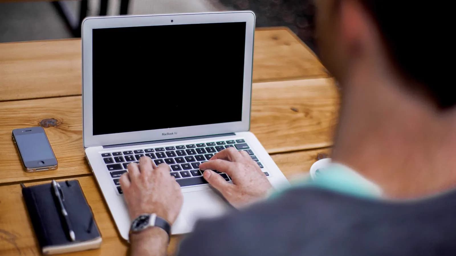 A person's hands typing on a laptop at a wooden desk, with a phone and notebook alongside