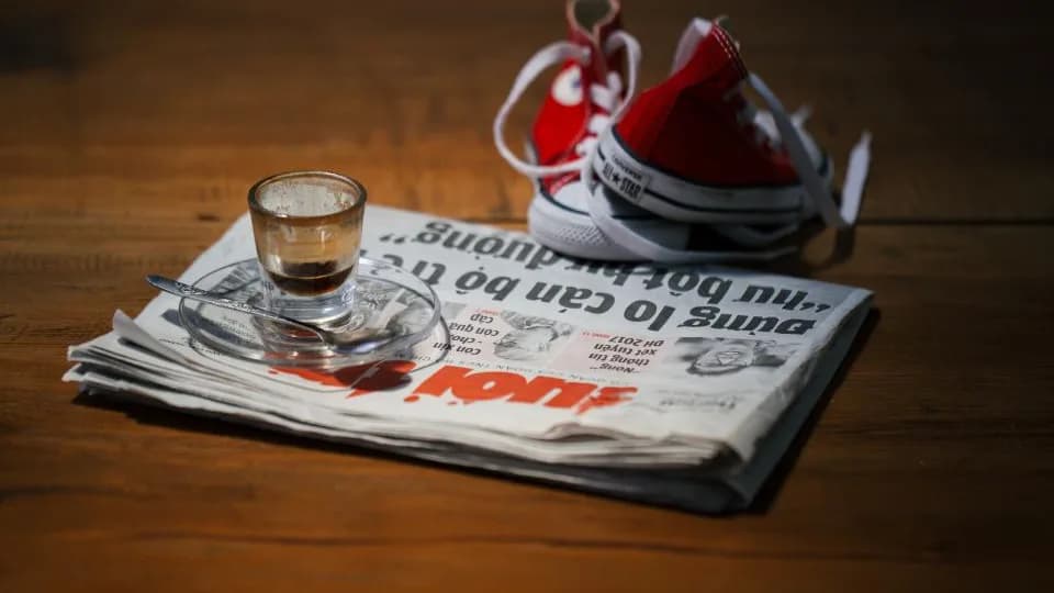 A folded newspaper on a wooden table with a cup of coffee and a pair of red shoes resting on it