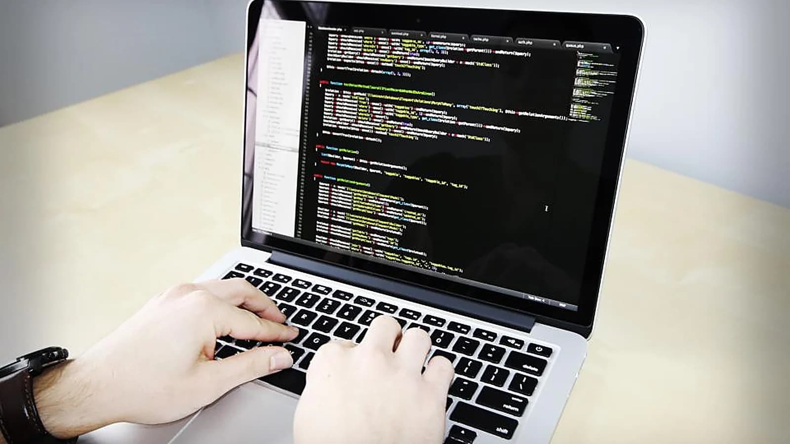 Photograph: two hands typing on a laptop at a pale desk, the screen filled with a syntax-highlighted code editor and a row of open file tabs