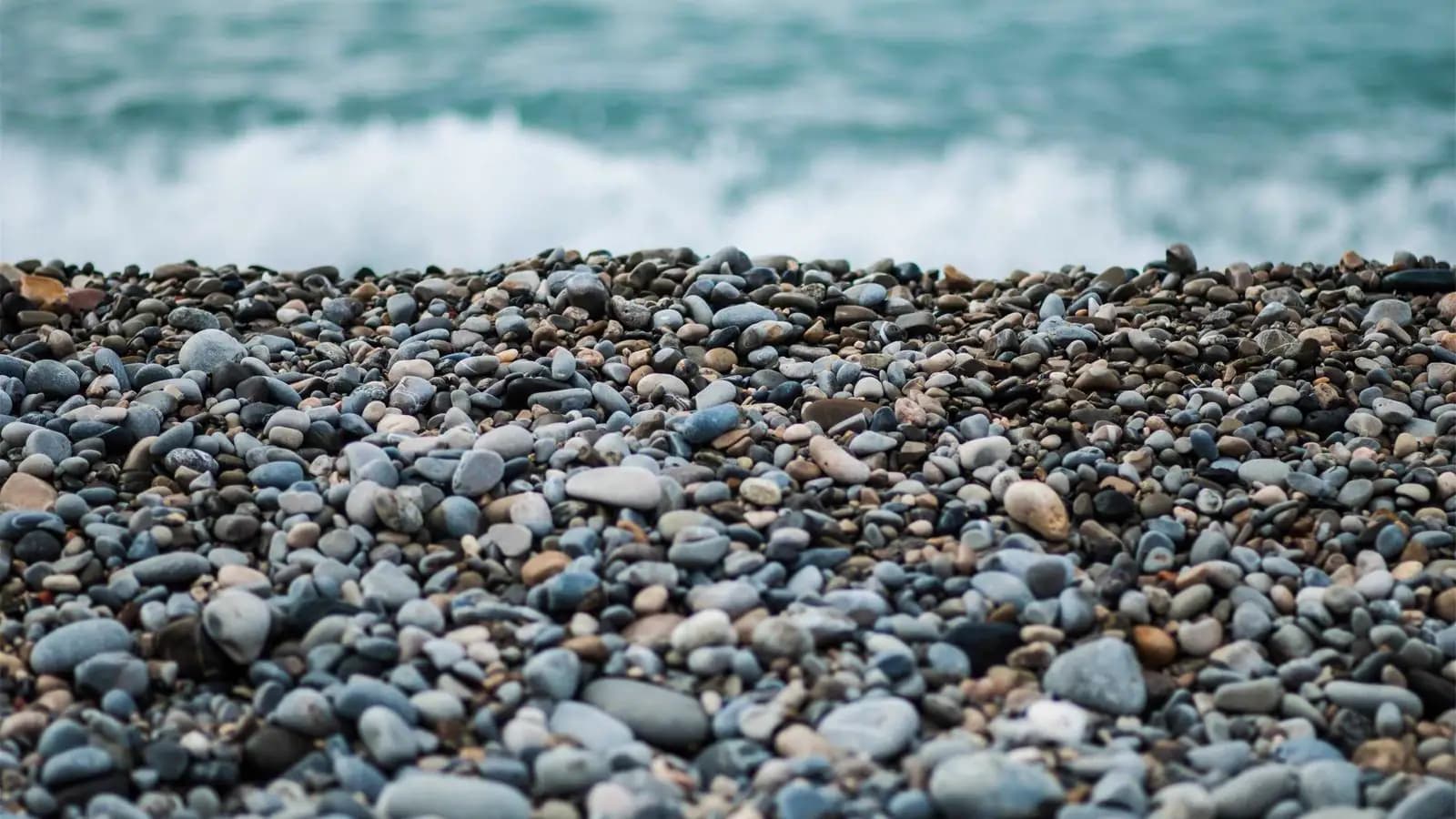 Smooth grey and blue pebbles on a beach with sea foam behind them