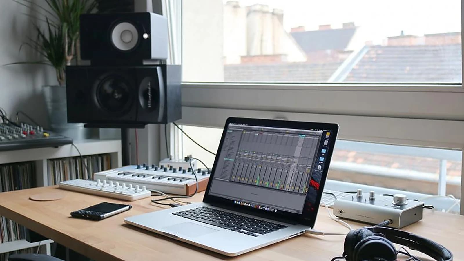 Photograph: a laptop on a desk by a window showing a multitrack recording timeline, with a mixer, a monitor speaker and headphones around it