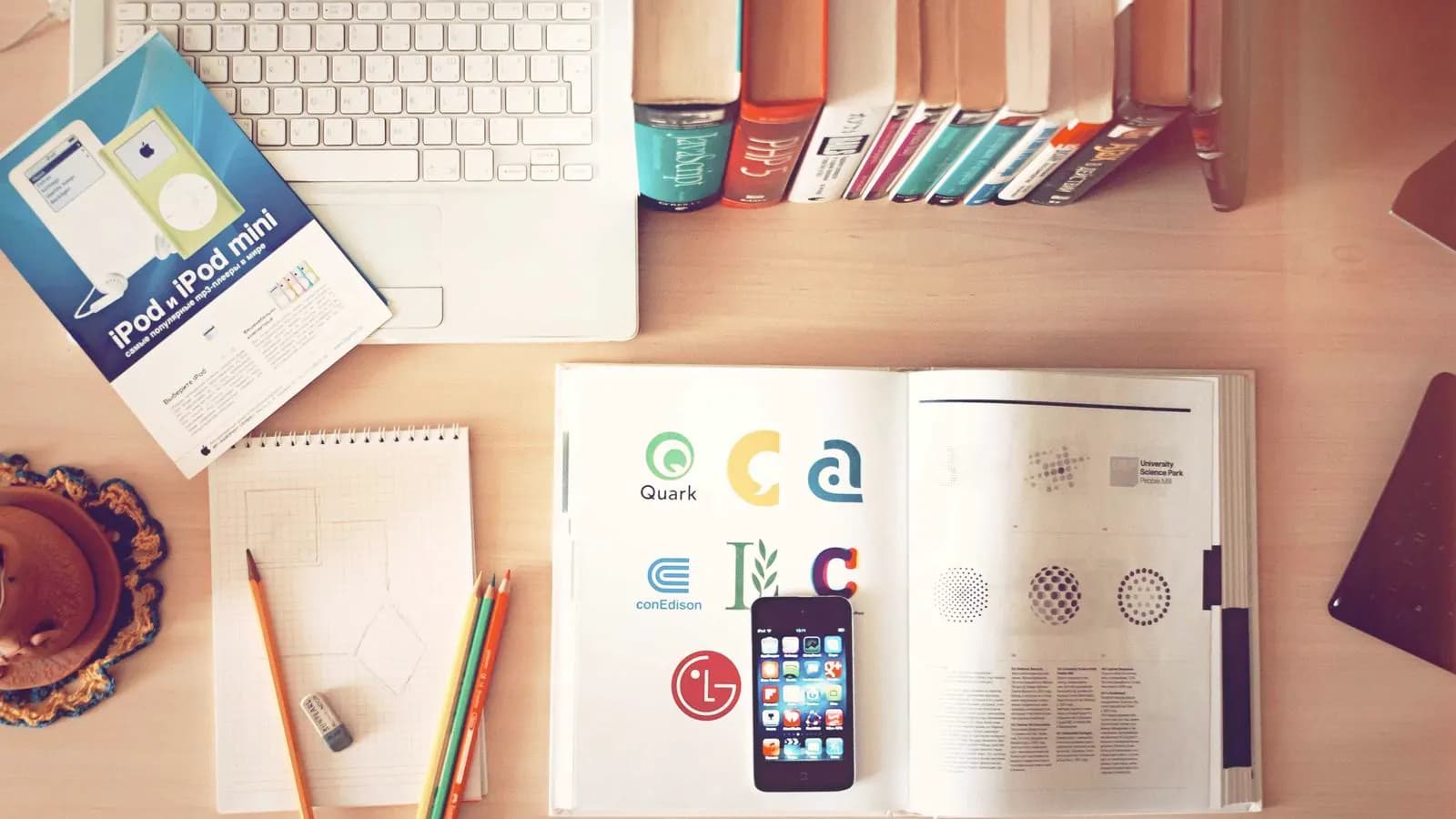 A desk seen from above with a keyboard, books, logo sketches, pencils and a phone