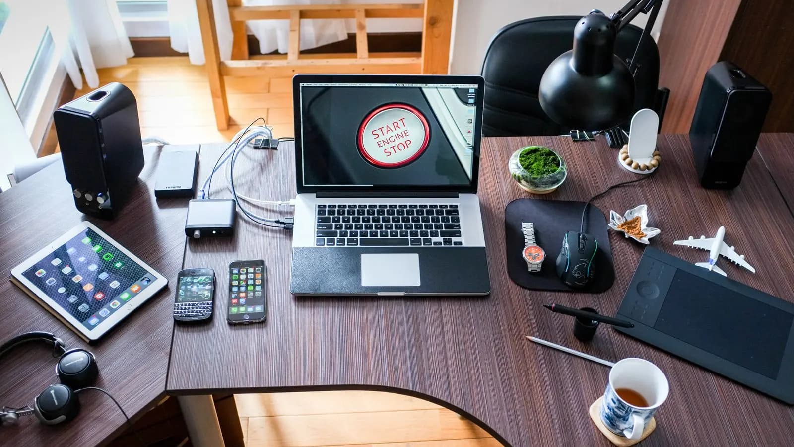 A busy wooden desk with an older laptop, a tablet, two phones, speakers and a coffee cup