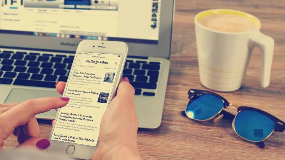 A hand holding a phone open on a news article, resting on a laptop keyboard beside a mug of coffee